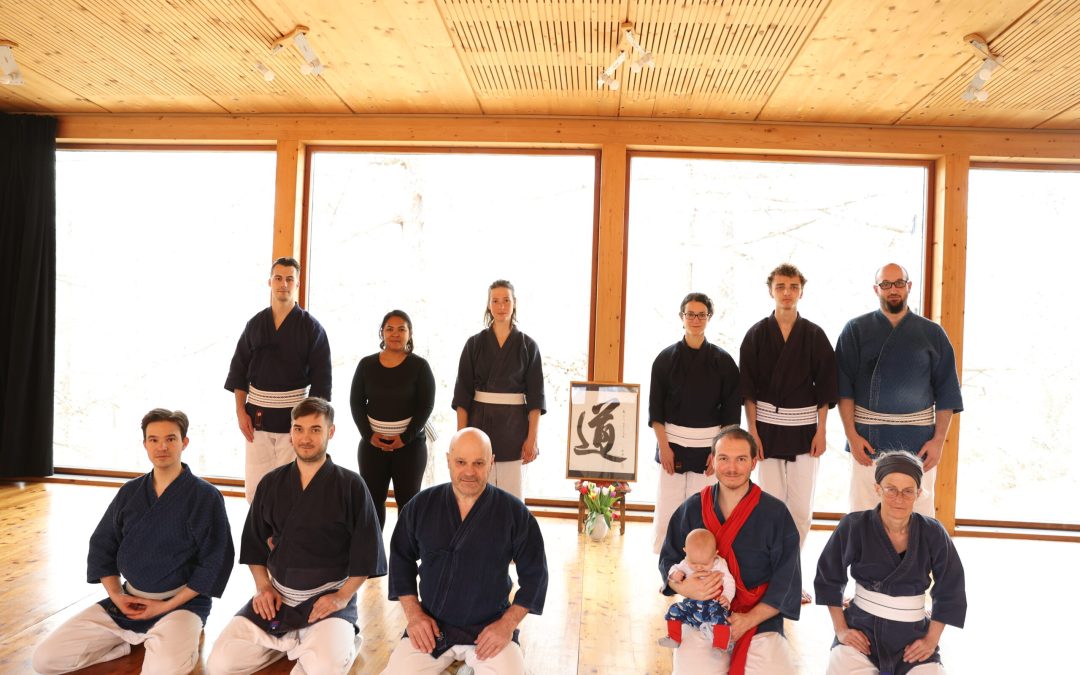 Photo de groupe à la fin du stage : tous les pratiquants posent ensemble devant le kamiza du dojo, les branches de mélèzes et la calligraphie « Dô » visibles à travers la vitre, ambiance chaleureuse et esprit d’unité.